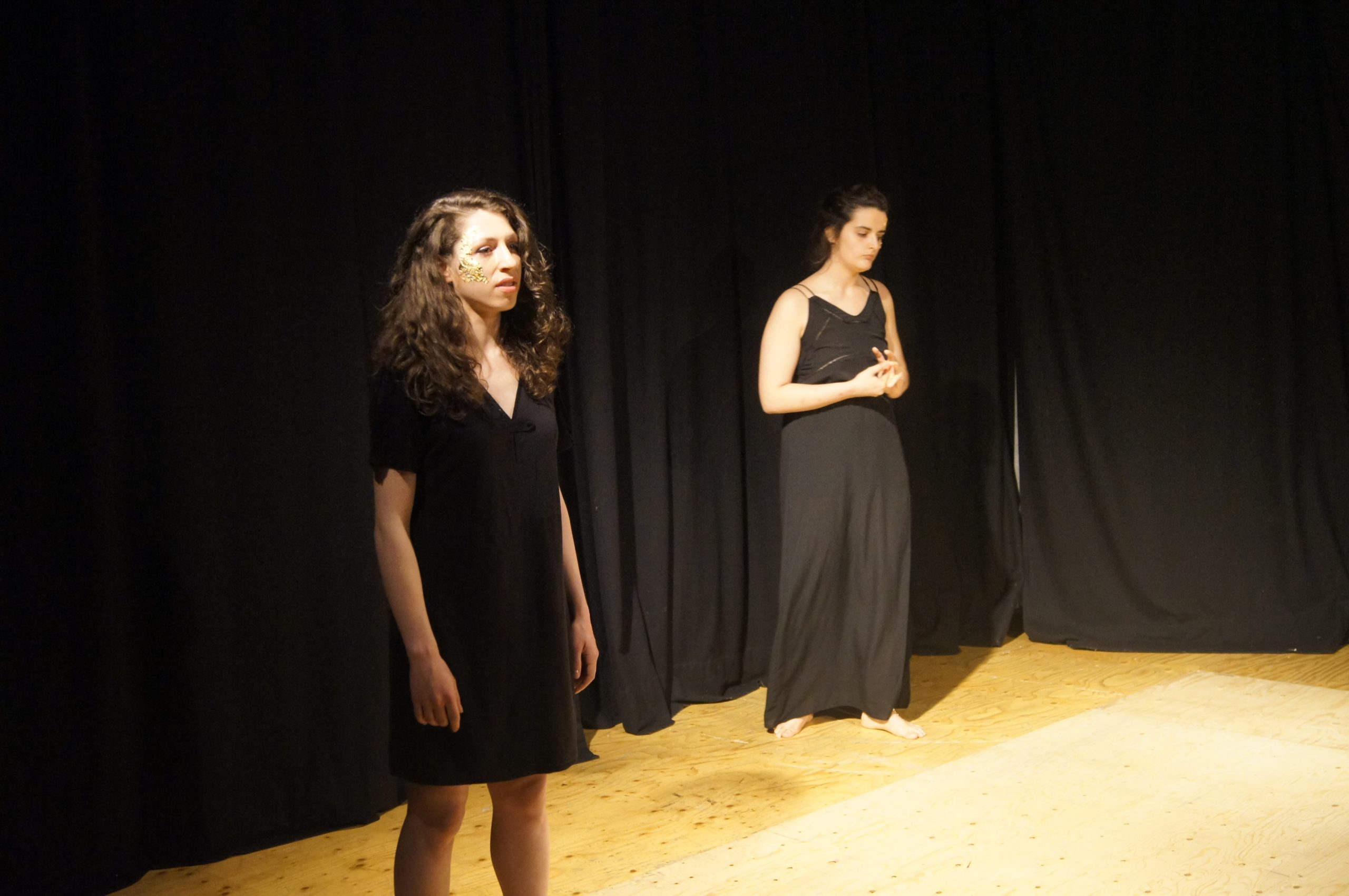 Theatre performance, two women on stage with black dresses. One is reciting, the other one looks sad or concerned