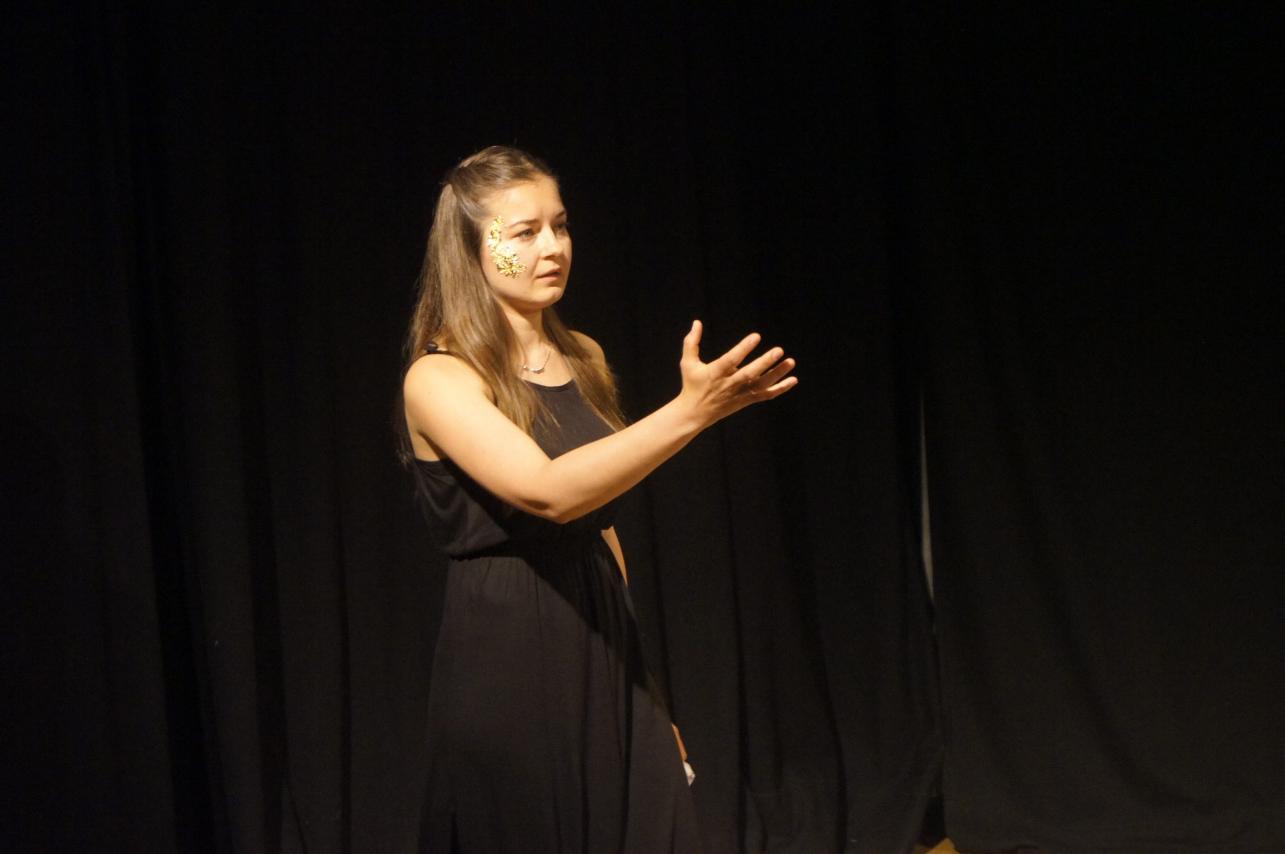 Photo from the theatre performance Keeping up with the Greeks. woman on stage with a black dress, dark blonde hair, golden flakes on her right cheek. Black curtain behind her. She is reciting