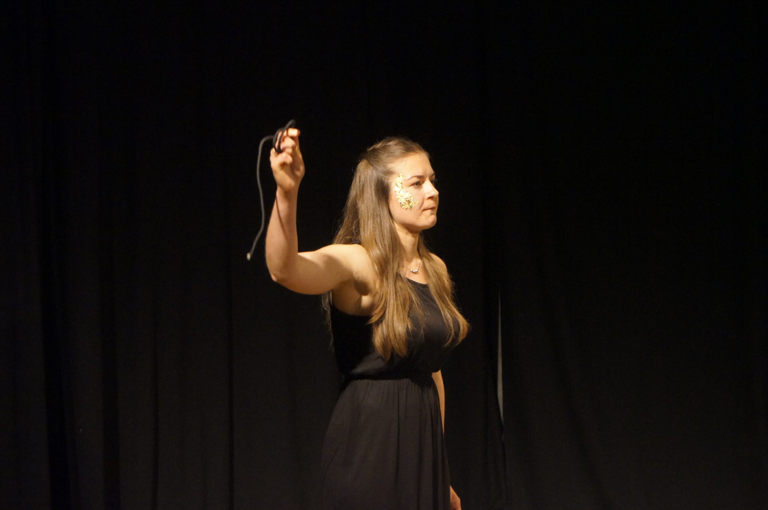 Theatre performance, woman on stage with a black dress, dark blonde hair, golden flakes on her right cheek, holding a string. Black curtain behind her. She looks angry