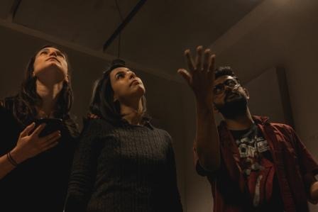 three actors looking up and beyond the photo. One of them is pointing with his arm open. One man and two women