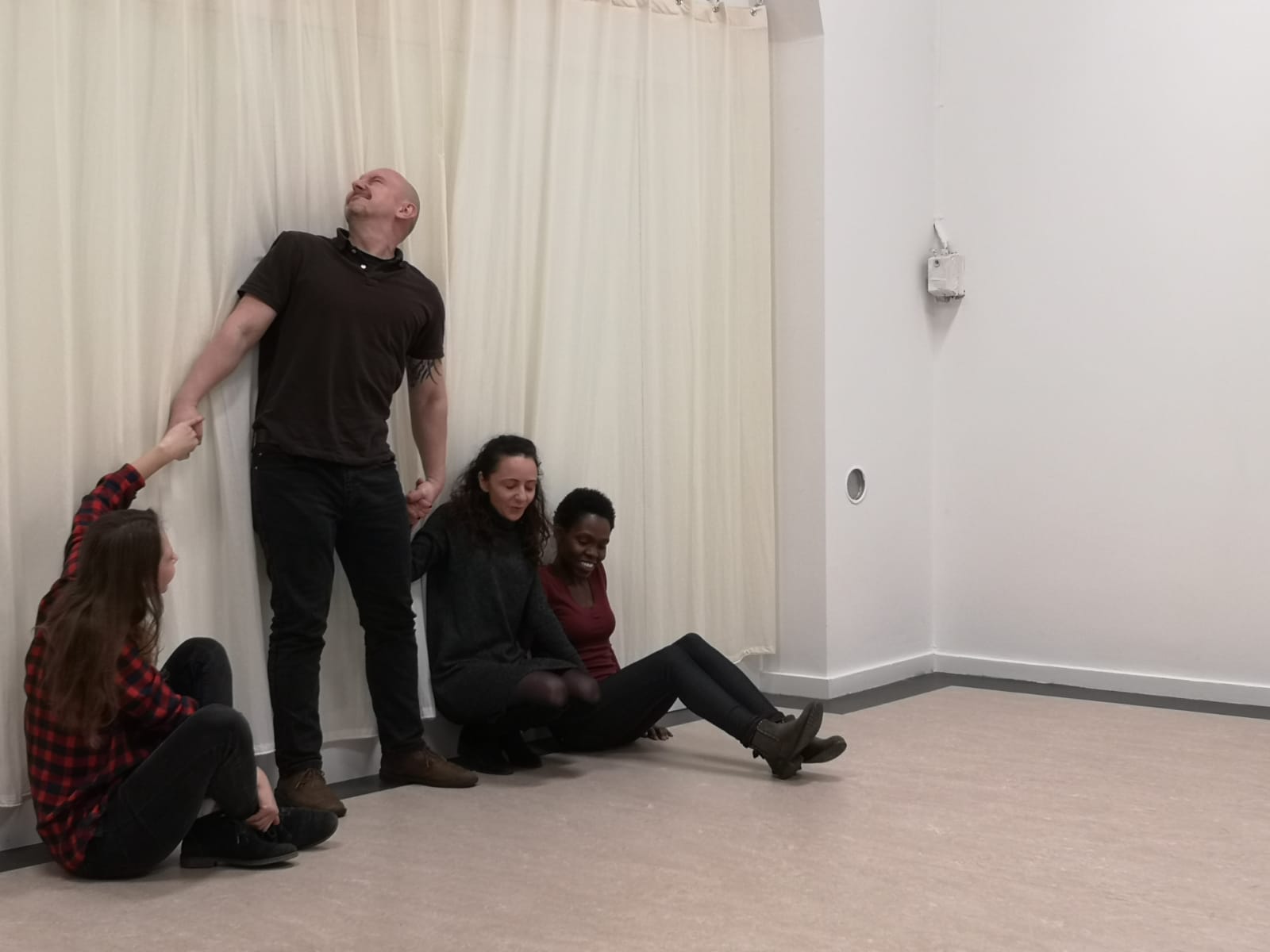 Acting Class Act Attack. Four people, three women, one man, are in a row and have their backs against the wall. The women sit on the floor. Two of them hold hands with the man who is standing