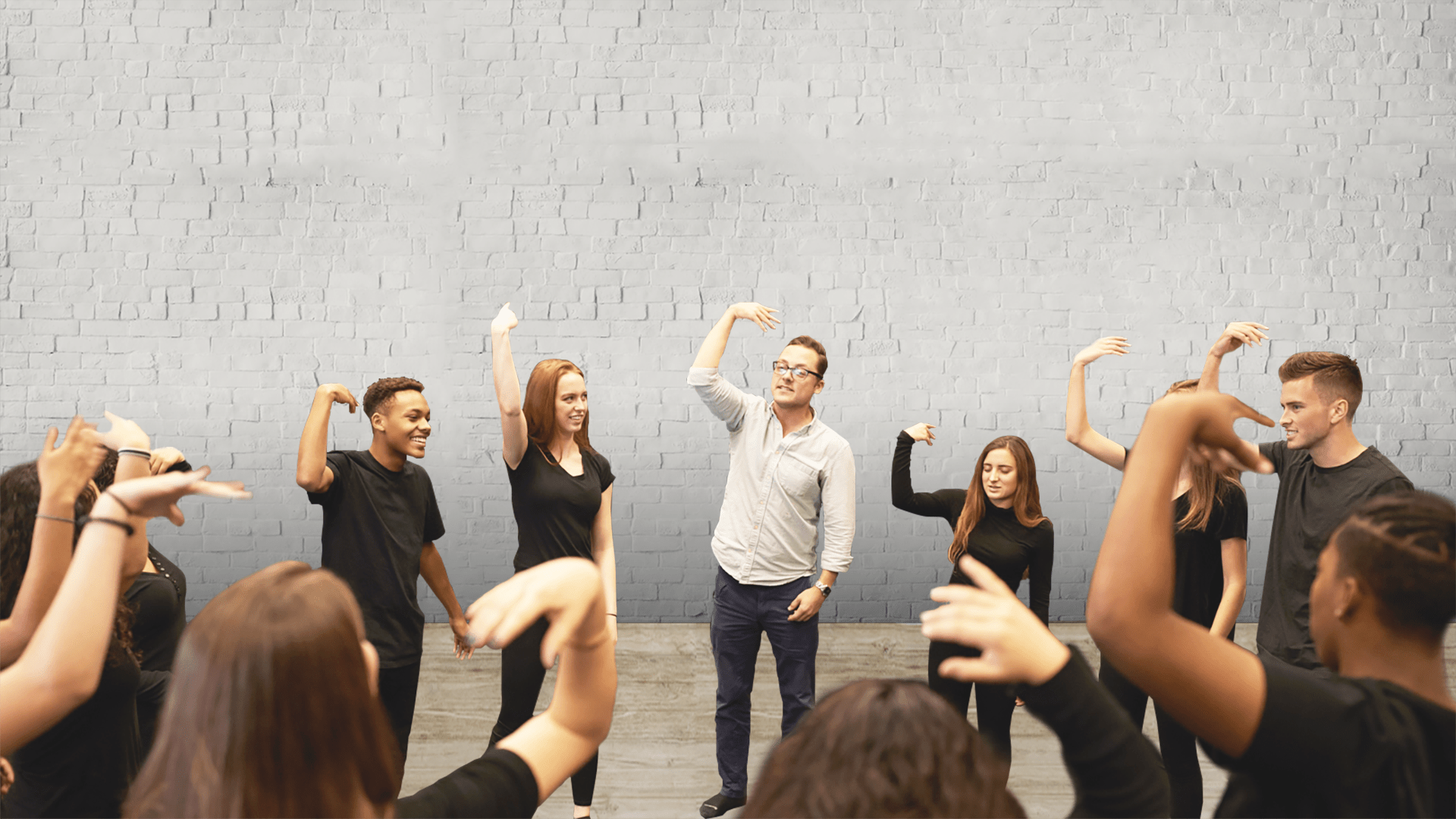 Actors practicing. A group of people wearing black, except for one man with glasses wearing a white shirt, have their arms lifted in a 90 degree angle with their wrist also in a 90 degree angle. Background: Grey brick wall
