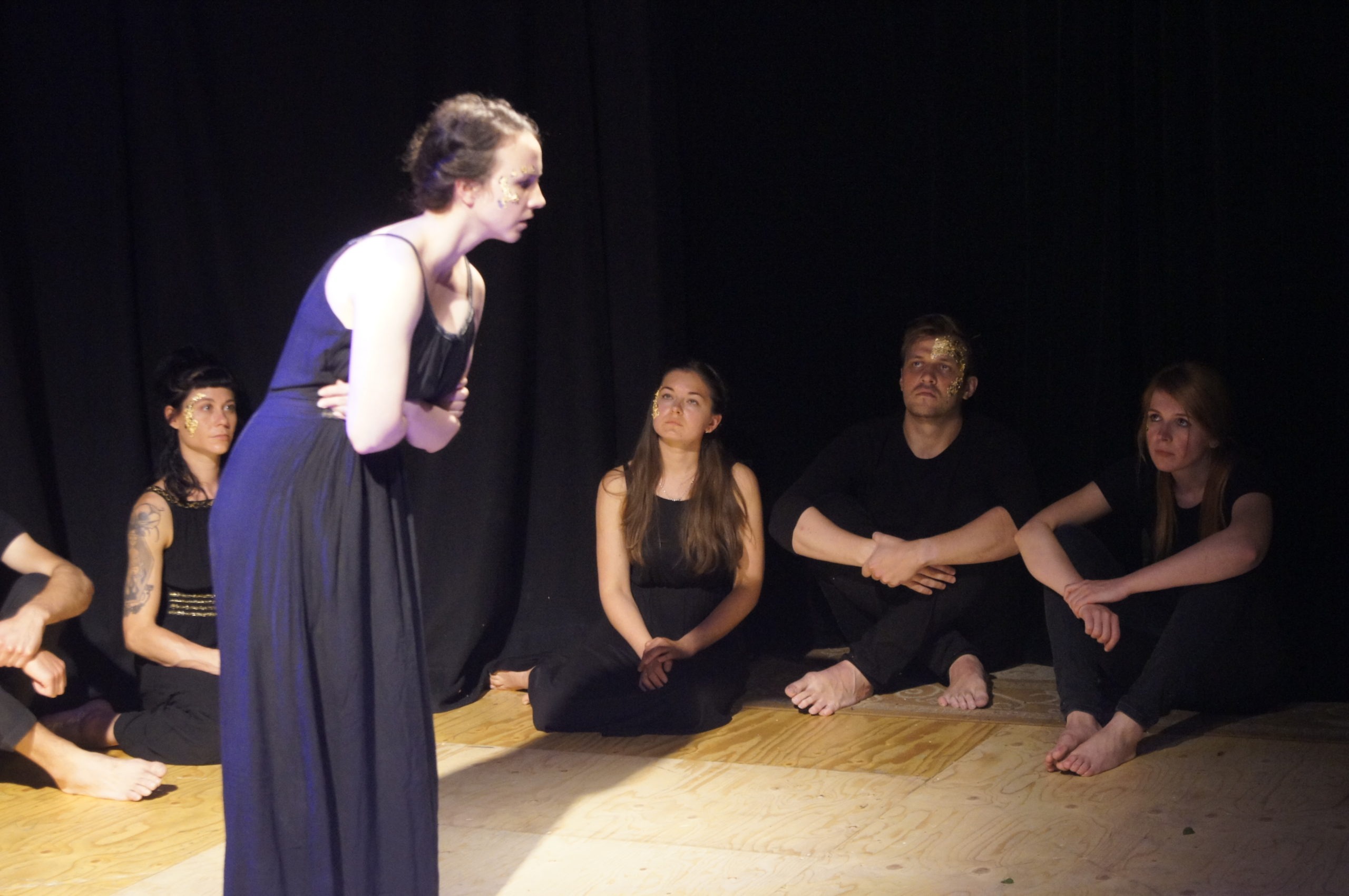 Photo from the theatre performance Keeping up with the Greeks. Woman on stage with a black dress, brown hair, golden flakes on her right cheek, holding her stomach. Behind her, four actors on the floor, black clothes, golden flakes on their face