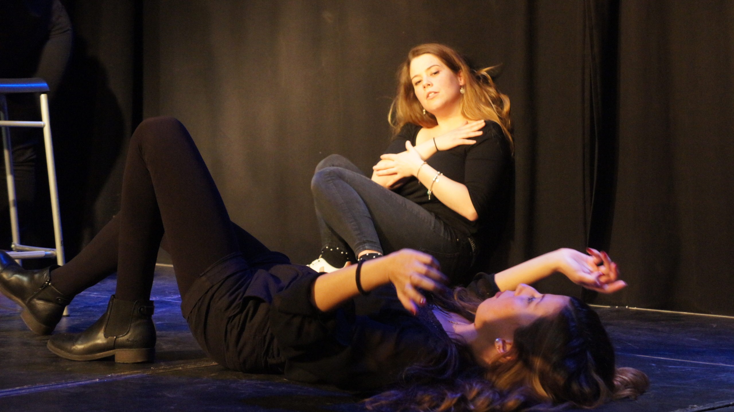 Act Attack's improv and physical show. Two girls on stage wearing black. One is laying on the floor, the other is sitting against a wall.