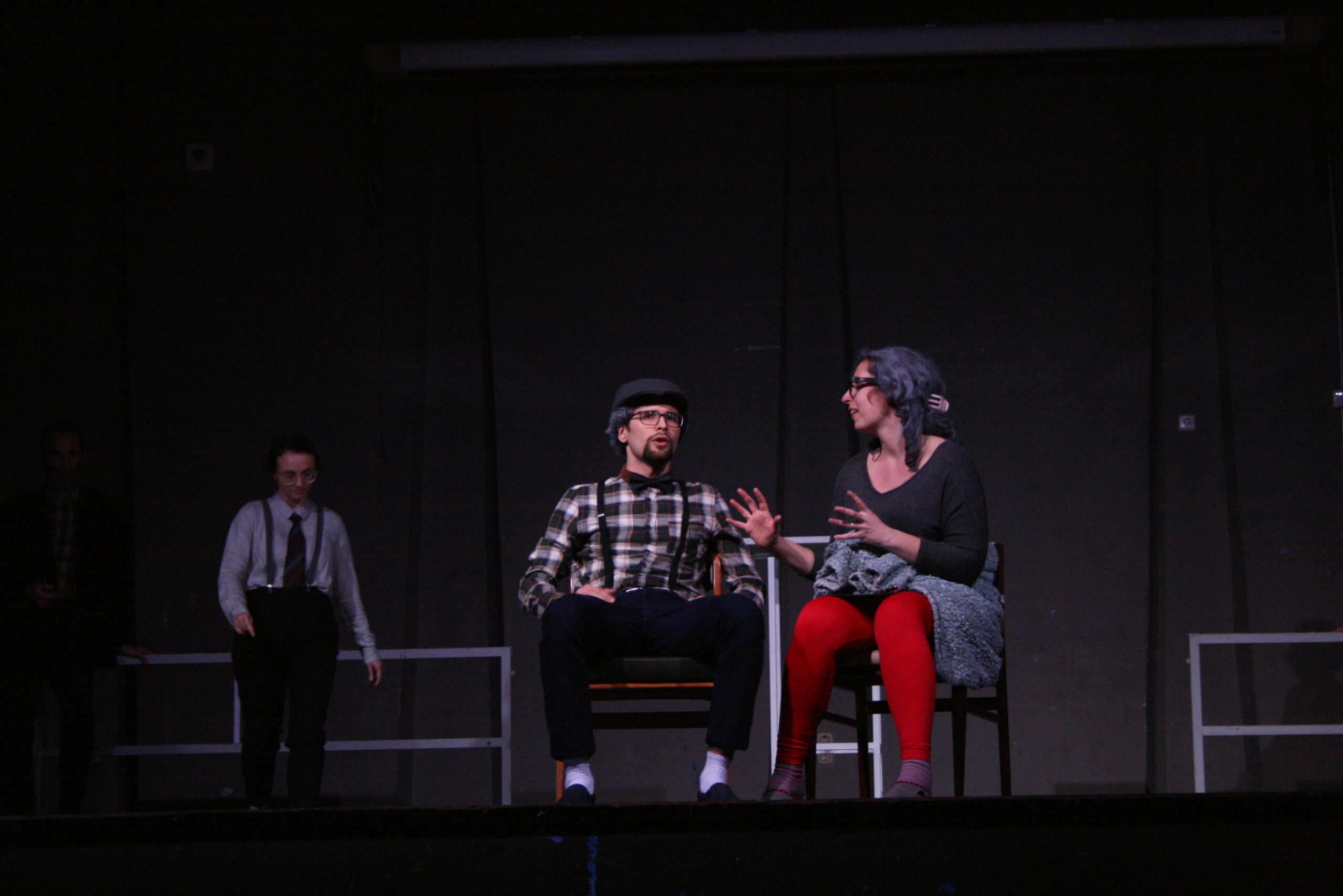 Photo from the show "The Chairs". A male and a female actor sit on chairs, talking. He looks over the audience and she looks at him, her arms open like holding a ball