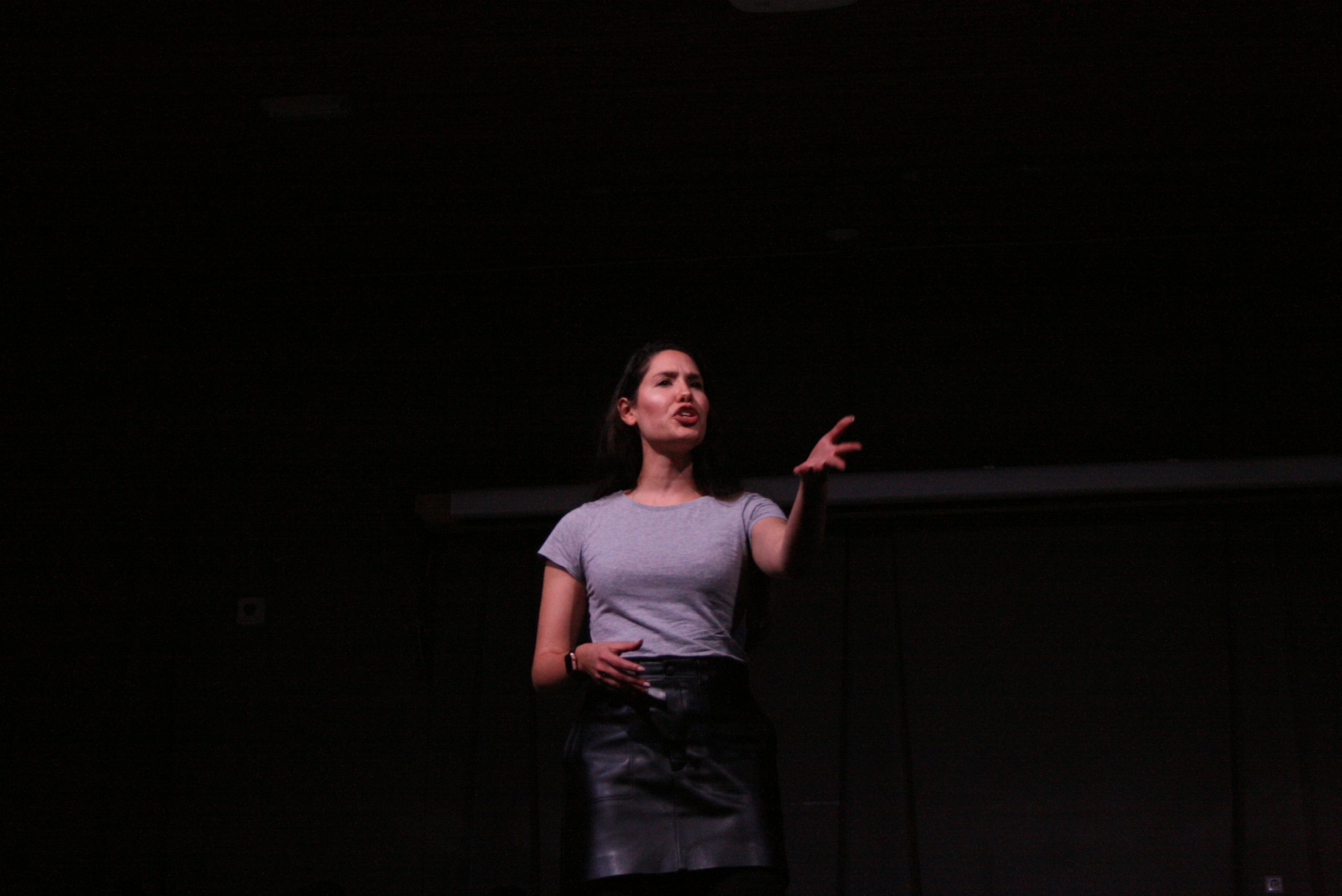Photo from the show "The Chairs". A female actor addresses the audience. She looks angry, reaching out one of her arms.