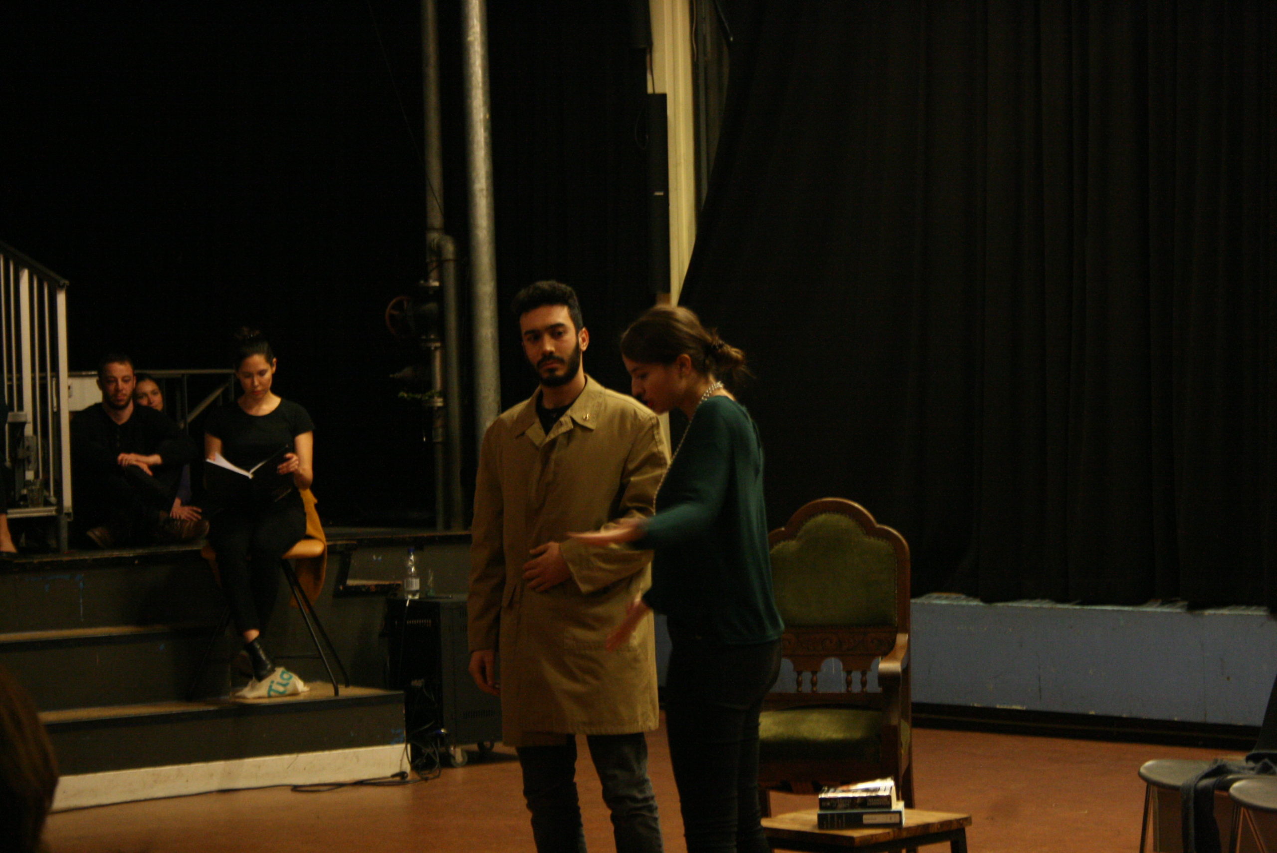 Photo from the show "Skin of our Teeth". Two actors are on stage, one dressed like a 1920s madame speaking to a man wearing a beige half-long coat