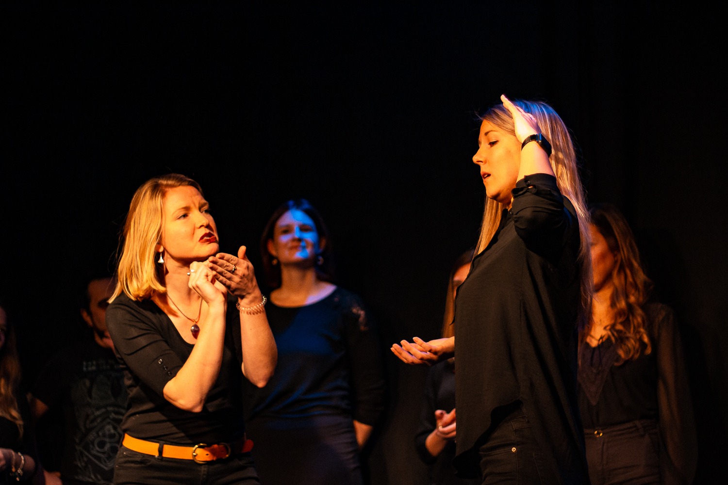 Act Attack's improv and physical show. A group of people on stage wearing black. Two blonde women talking or gesturing