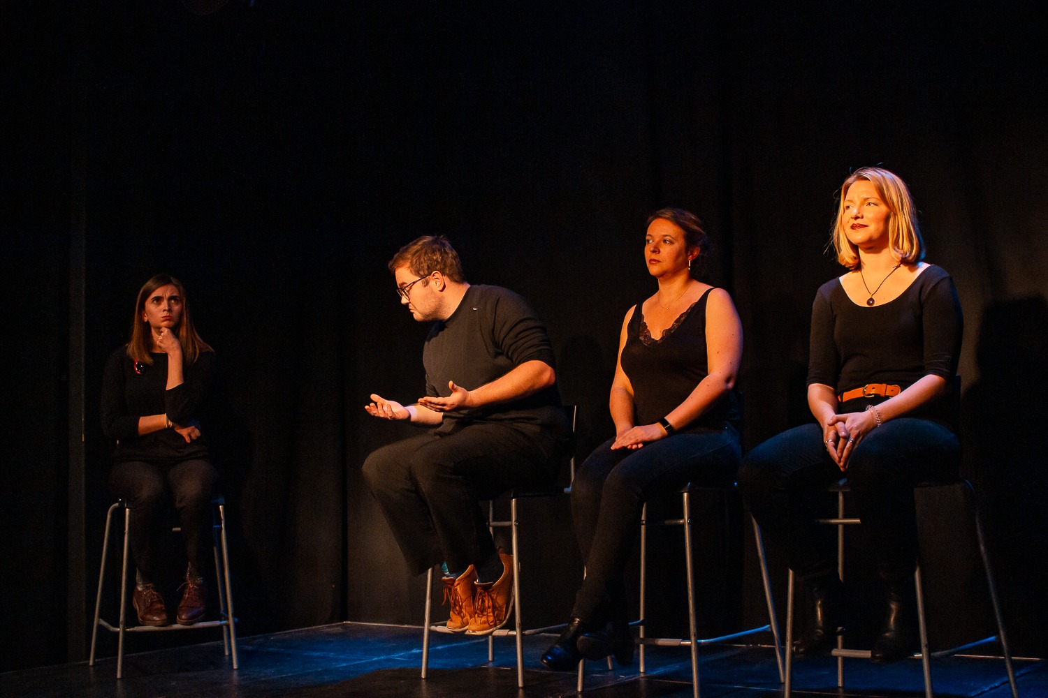 Act Attack's improv and physical show. A group of people on stage wearing black, sitting on bar stools. Three women and a man making a gesture like he's wondering